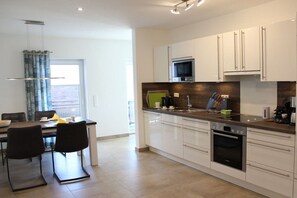 A modern kitchen with white cabinets, a microwave, a dishwasher, and a dining area with a table and chairs. - At the canal footbridge (Krummhörn-Greetsiel)
