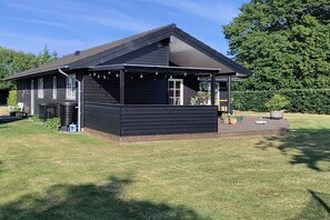 A black wooden building with a sloped roof, a covered porch, and a small garden area with potted plants. - Top luxury class on a dream beach (Marielyst)