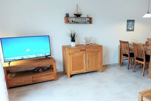 A living room with a TV, wooden cabinet, dining table, chairs, and a small shelf with decorative items. - Holiday home with 1 bedroom (Dornumersiel)