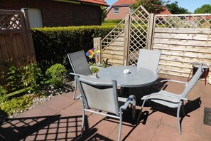 Eine Terrasse mit einem runden Tisch und vier Stühlen, umgeben von einem Holzzaun und einem Heckenstreifen.
