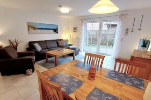 A living room with a sofa, a dining table with chairs, a coffee table, and a large window with curtains. - Holiday home with 1 bedroom (Dornumersiel)