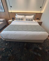 Habitación de hotel con una cama grande, una mesita de noche con lámpara, una silla y un televisor montado en la pared.