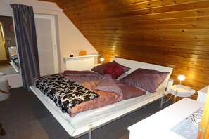 Een slaapkamer met een houten plafond, een bed met veel kussens, een nachtkastje met een lamp en een radiator.
