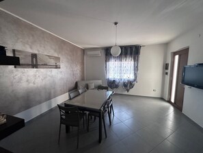 A modern living room with a dining area, a flat-screen TV mounted on the wall, and a large window with curtains. - Holiday Home 'Angel’s House' (San Giovanni Suergiu)