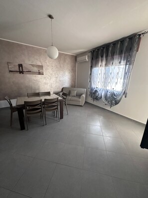 A modern living room with a dining area, a sofa, a table with chairs, a pendant light, and a window with sheer curtains. - Holiday Home 'Angel’s House' (San Giovanni Suergiu)