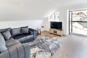 Una sala de estar moderna con un sofá gris, un televisor de pantalla plana sobre un mueble de madera y un gran ventanal con vistas a edificios.