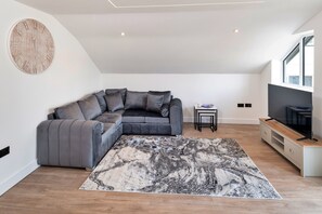 A modern living room with a sectional sofa, a marble-patterned rug, a TV on a wooden stand, and a large window providing natural light. - Finest Retreats | Greenswood (Brixham)