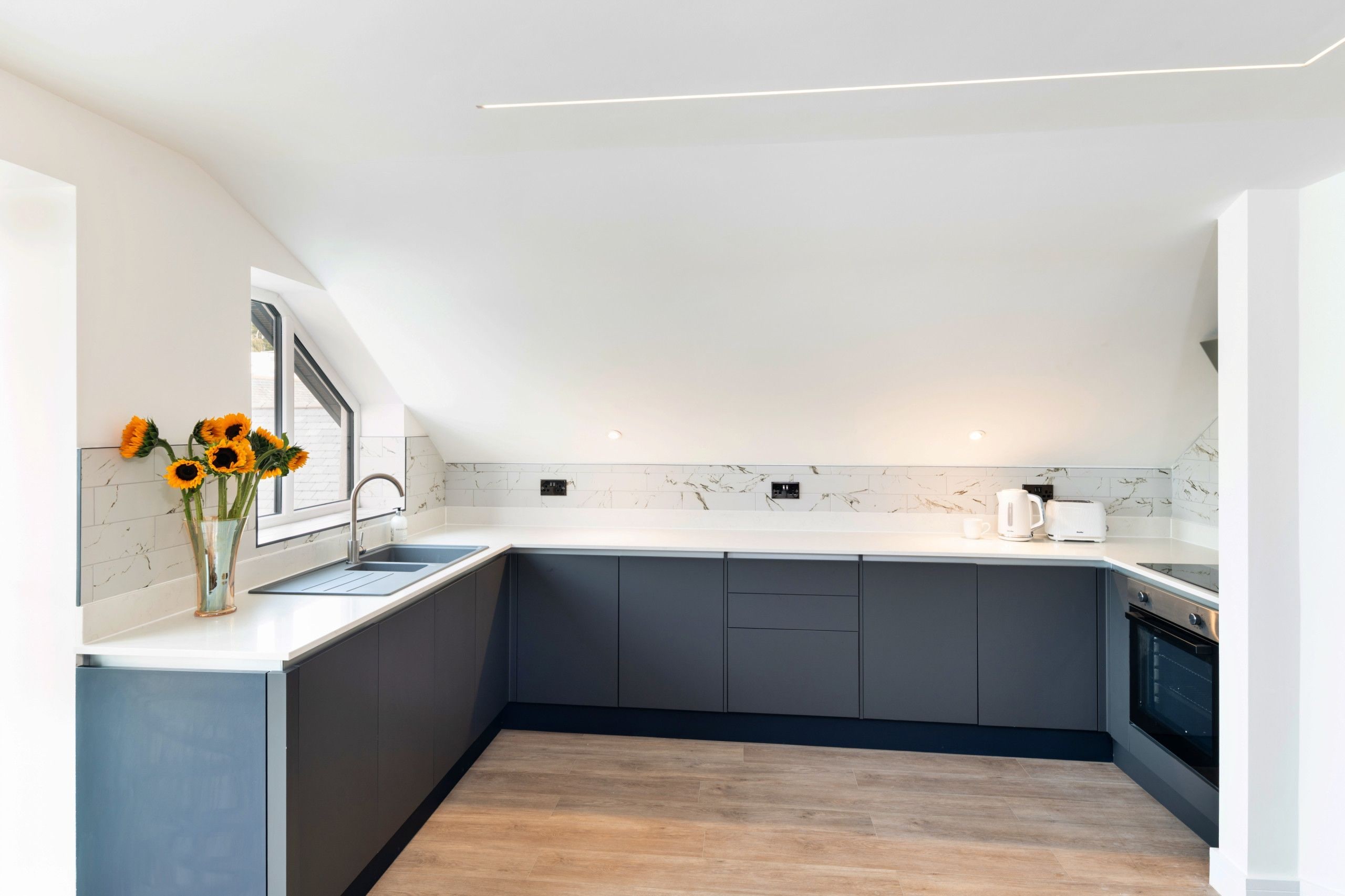 A modern kitchen with a skylight, dark cabinets, white marble backsplash, and a wooden floor.