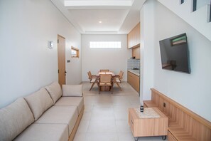 A modern living room with a sofa, a dining table with chairs, a TV mounted on the wall, and a kitchen area with cabinets and a stove. - Saura Villas (Buduk)