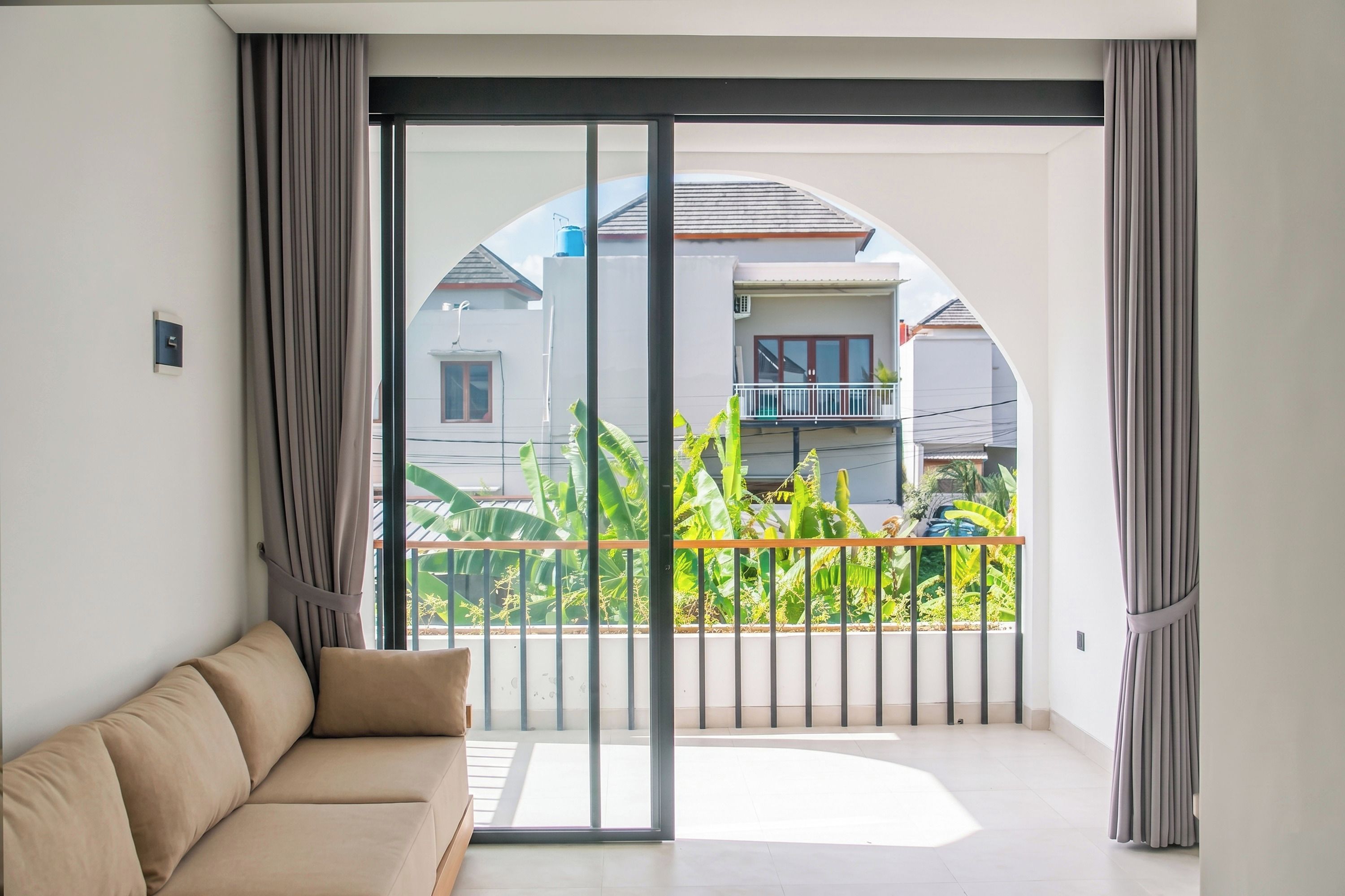 A modern living room with a sofa, large sliding glass doors leading to a balcony with a view of neighboring houses and greenery.