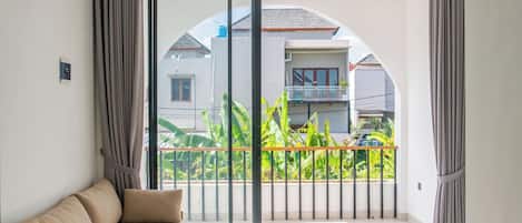 A modern living room with a sofa, large sliding glass doors leading to a balcony with a view of neighboring houses and greenery.