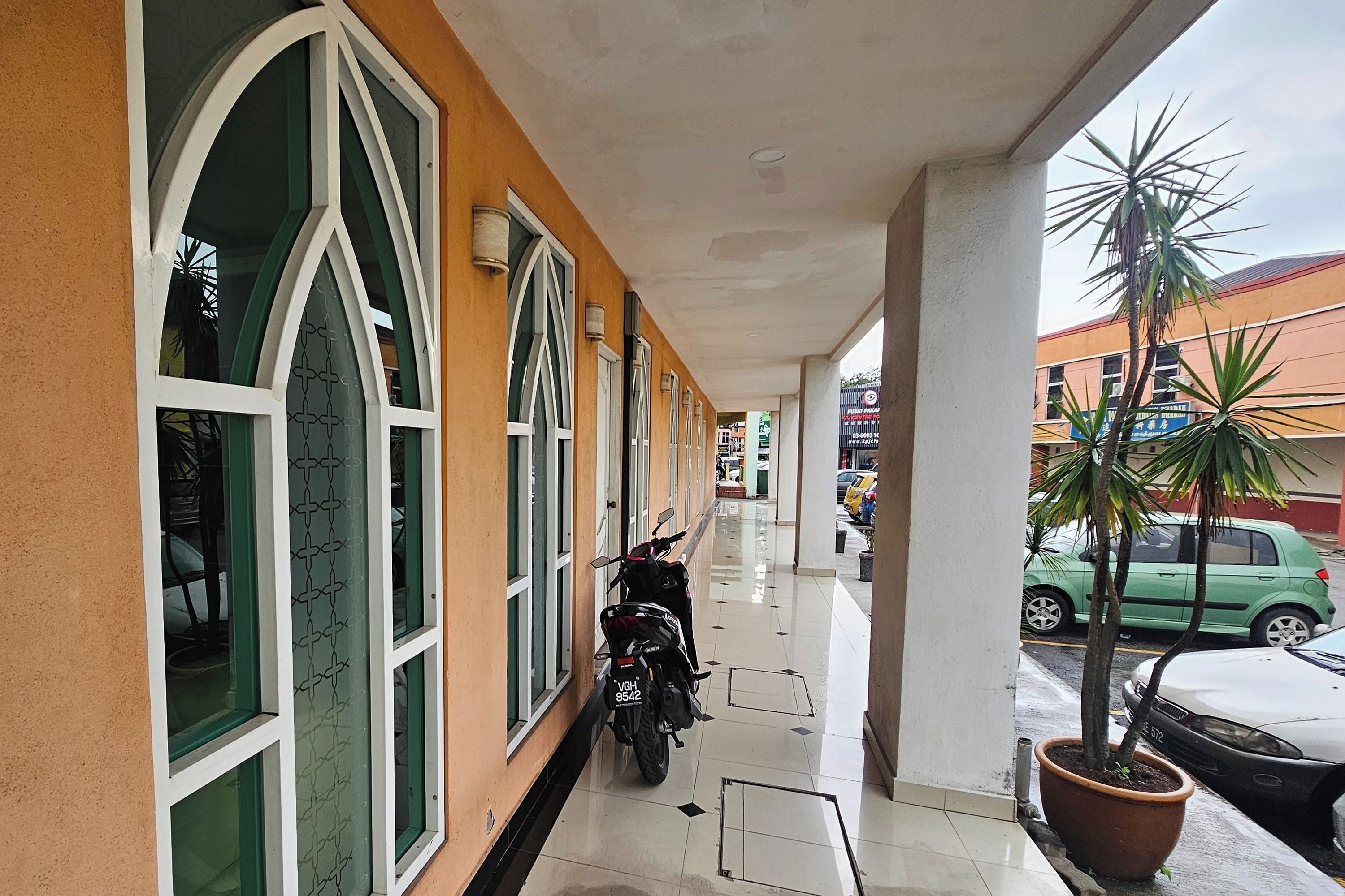 A covered walkway with a motorcycle parked, potted plants, and a green car parked outside.