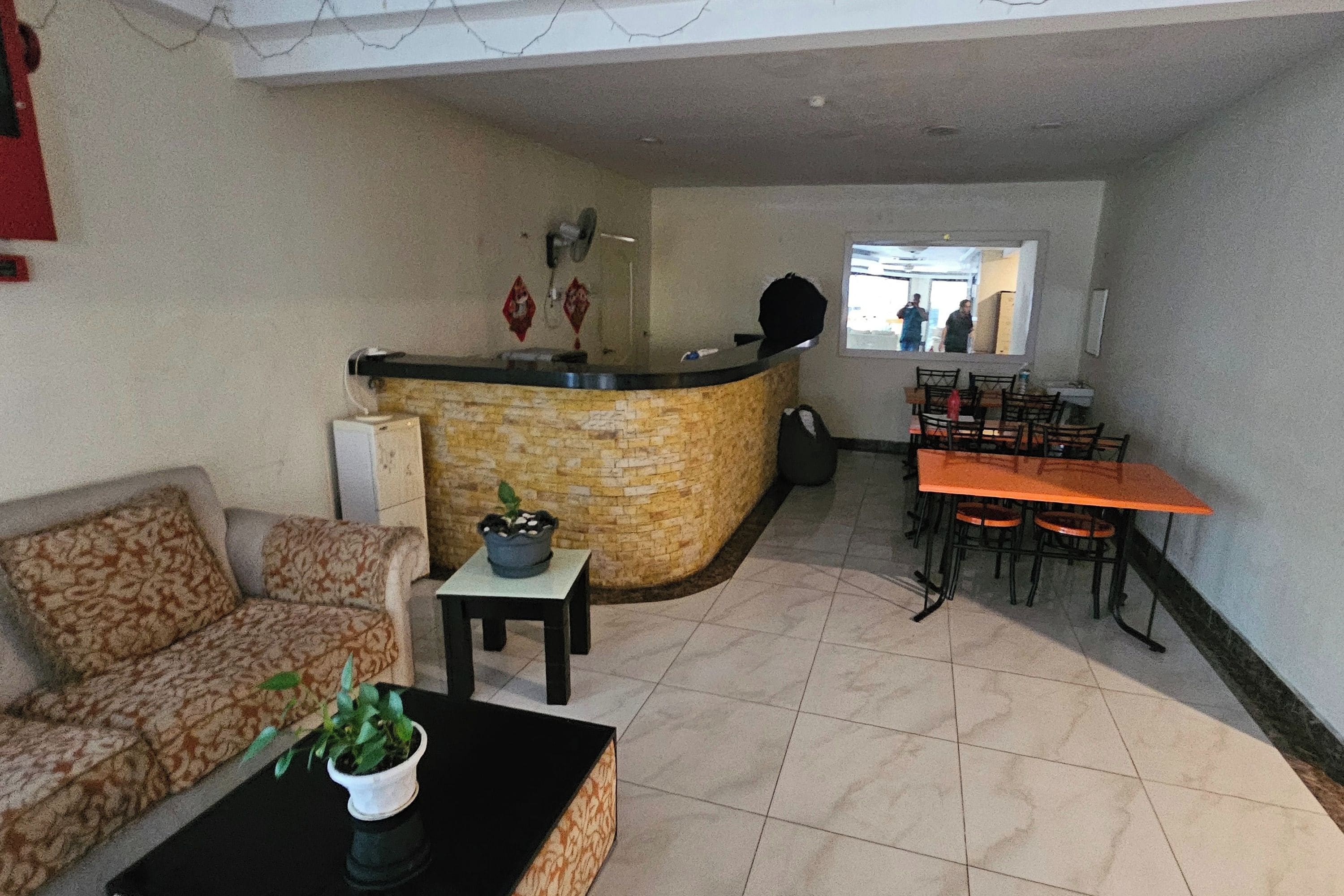 Una sala de estar con un sofá, una mesa de centro y una pequeña mesa con una planta. Se observa un mostrador de recepción con una silla y una ventana al fondo.
