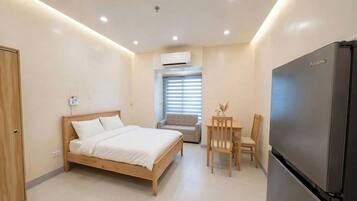 A compact bedroom with a bed, a dining table with chairs, a refrigerator, and an air conditioning unit.