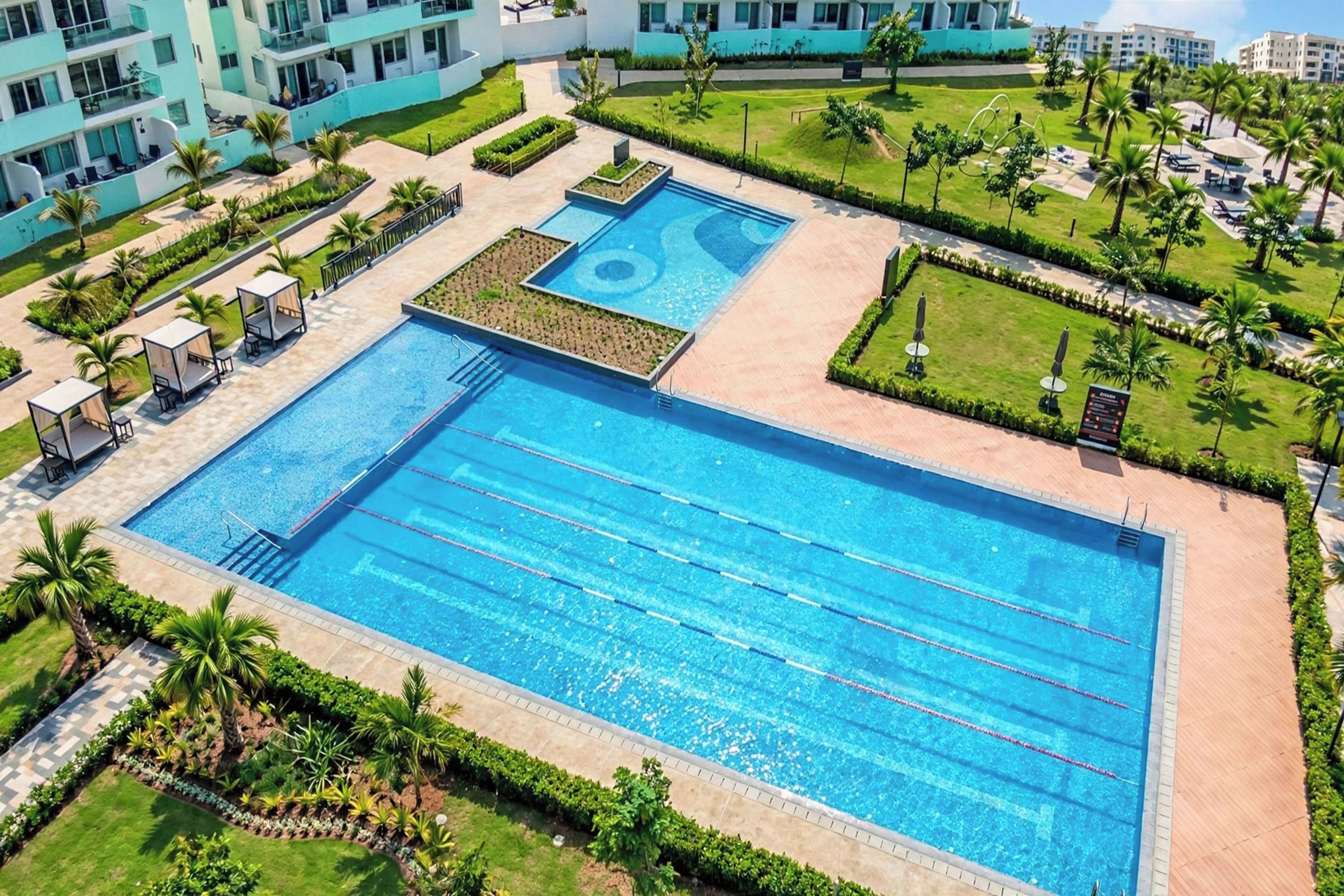 A large rectangular swimming pool with multiple lanes, surrounded by well-maintained lawns and palm trees.
