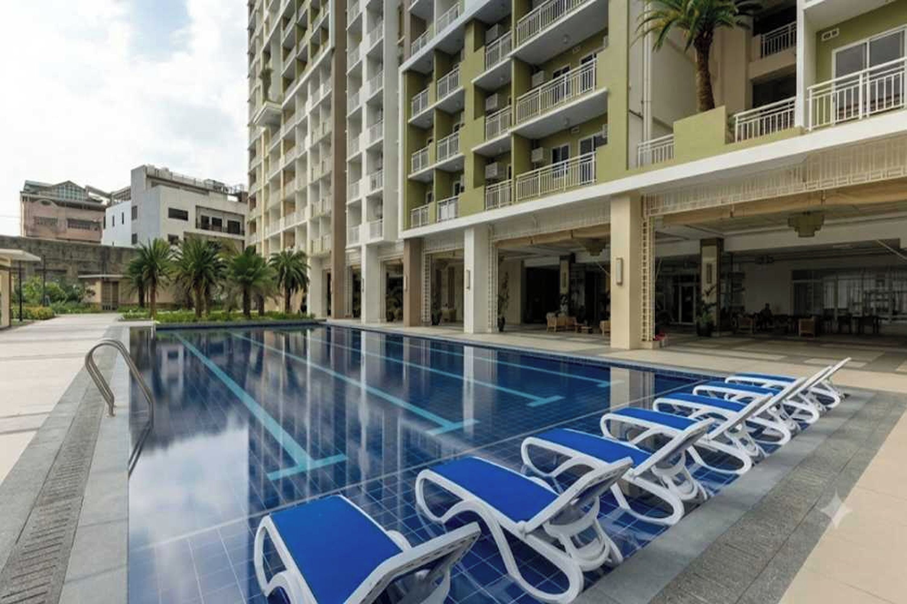 A swimming pool with lounge chairs, a multi-story building with balconies, and palm trees.