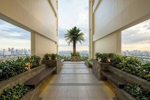 A corridor with a palm tree and plants, leading to a city view. - Cozy 1Br Staycation Unit At The Celandine, Balintawak, Quezon City (Quezon City)