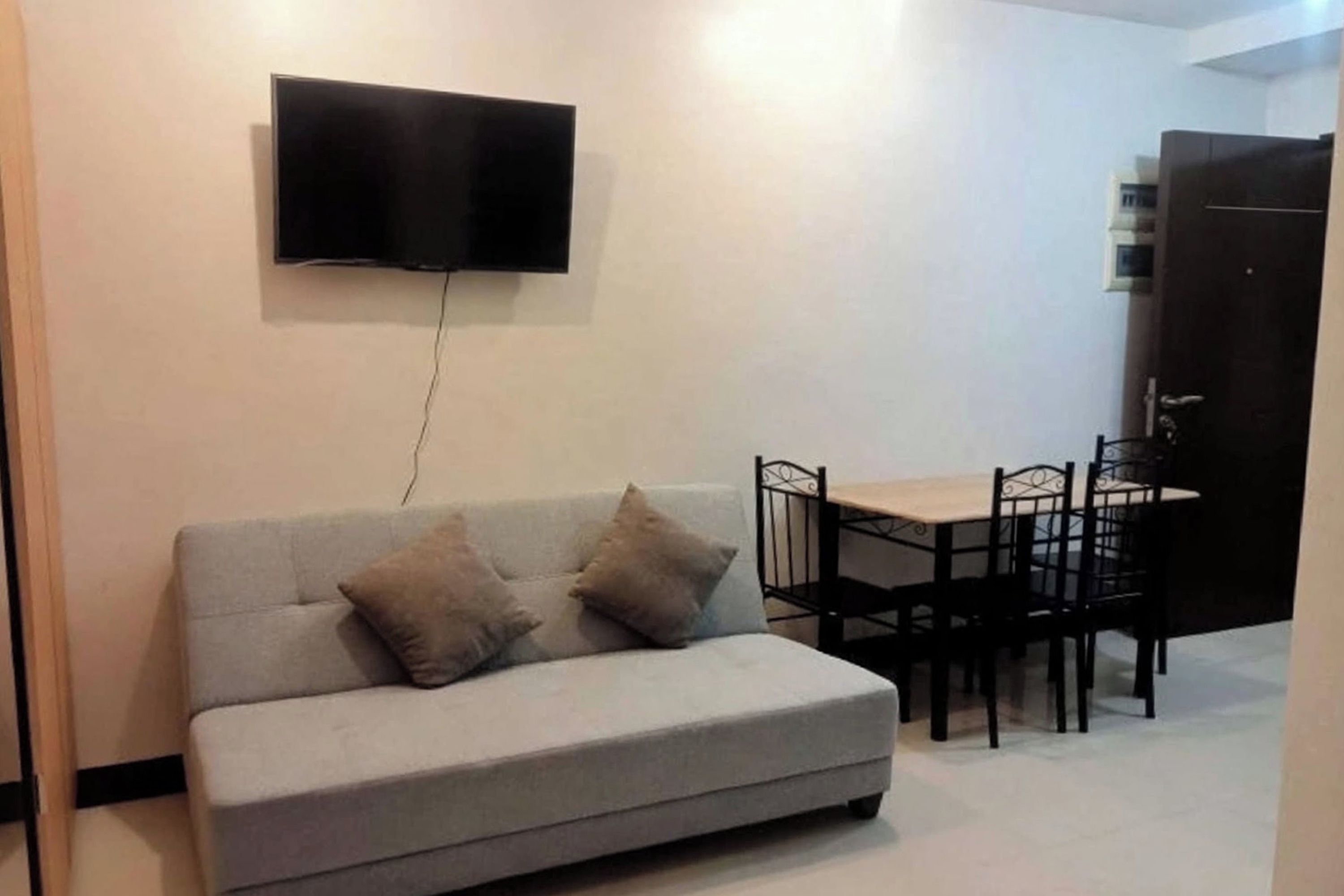 A living room with a grey sofa, two cushions, a flat-screen TV mounted on the wall, and a dining table with chairs.