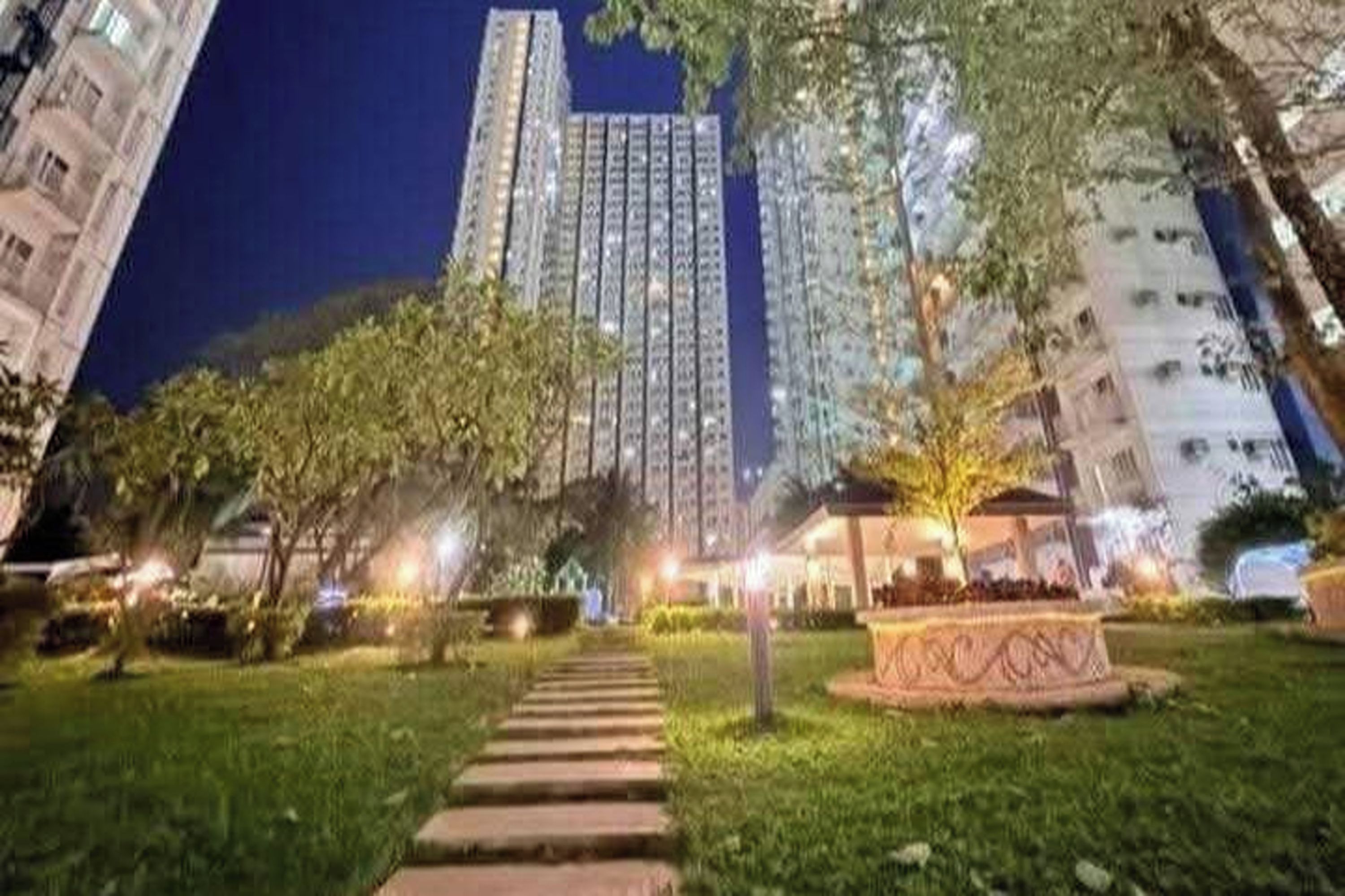 Vista noturna de um bairro residencial, com um caminho bem iluminado que leva a um edifício com vários andares.