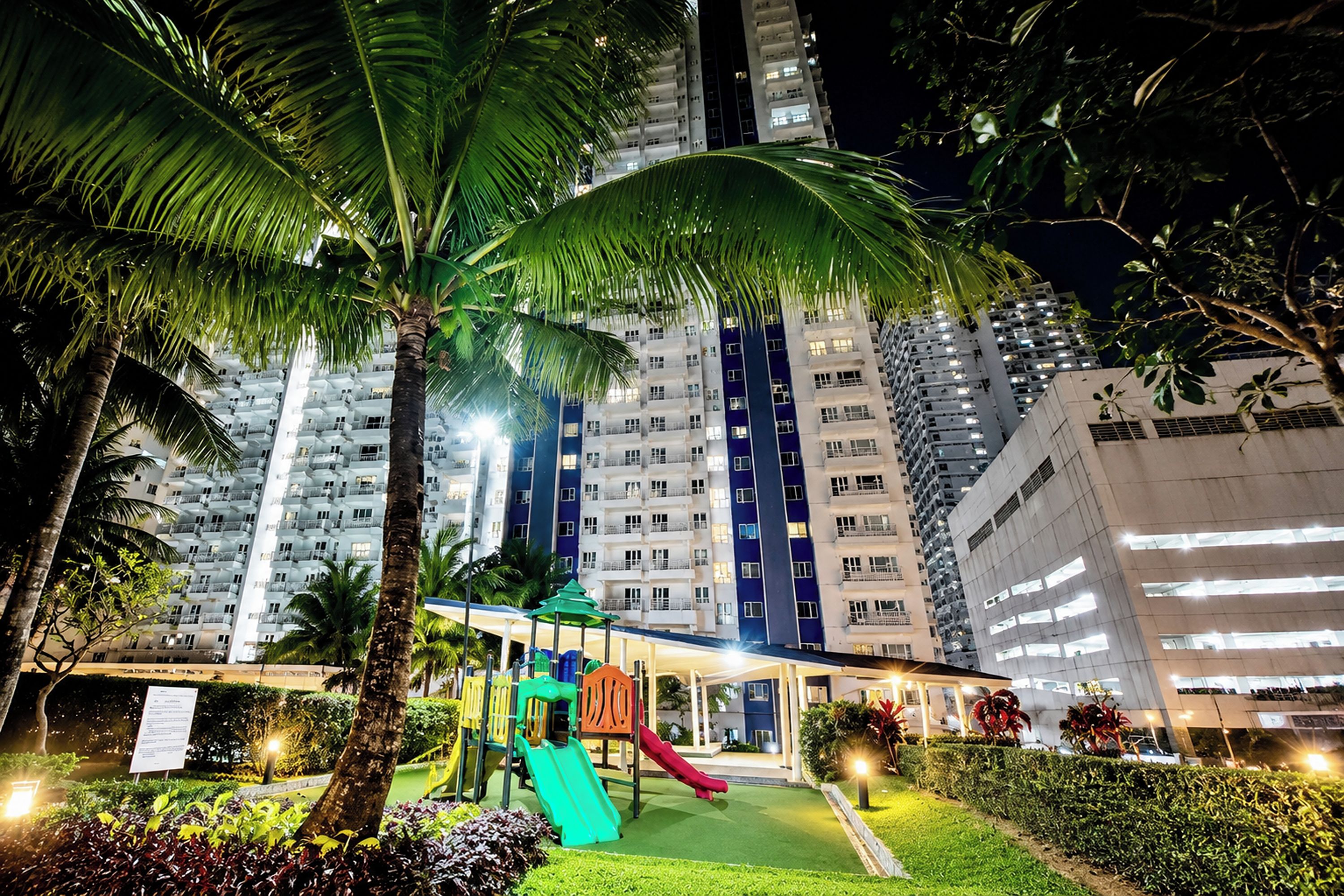 Un parque infantil con toboganes y estructuras para escalar, rodeado de palmeras y un edificio alto al fondo.