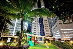 Un parque infantil con toboganes y estructuras para escalar, rodeado de palmeras y un edificio alto al fondo.