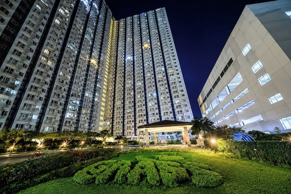 Un edificio de gran altura con varios pisos, un jardín bien cuidado con setos geométricos y un cielo nocturno despejado.
