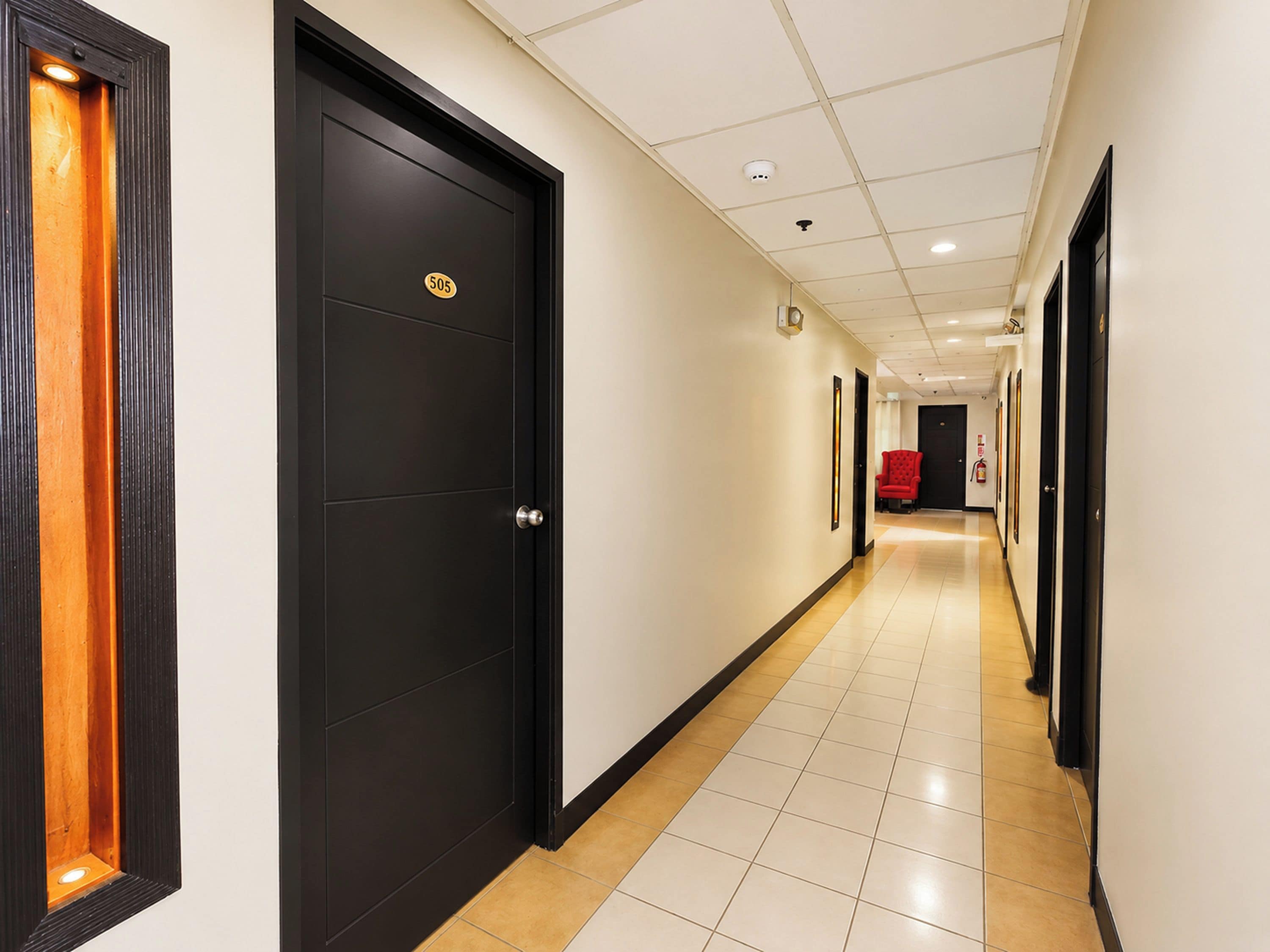 Corredor de hotel com portas pretas, piso de azulejos e uma cadeira vermelha ao fundo.