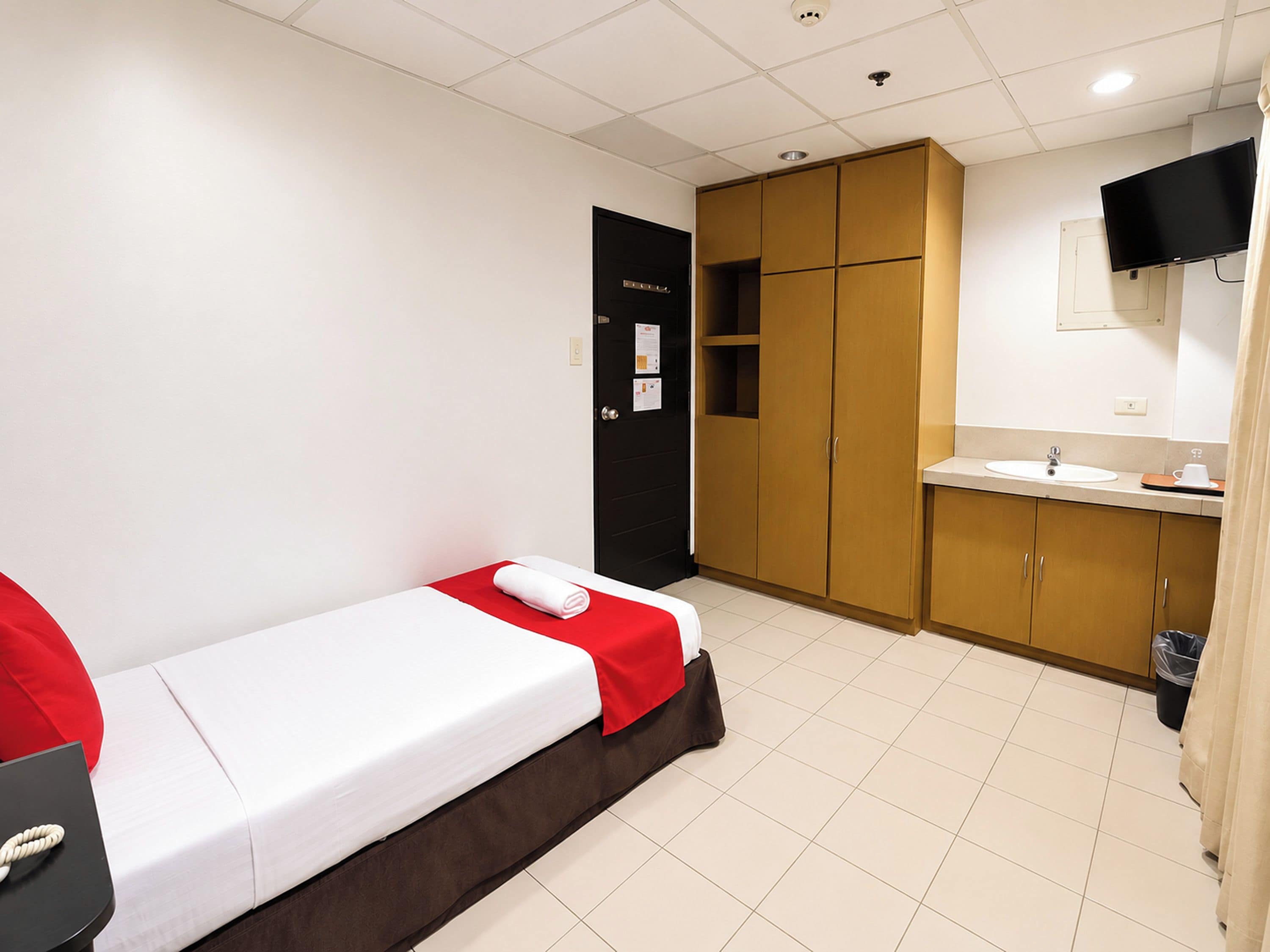 Habitación de hotel con una cama individual, un escritorio pequeño, un baño con lavamanos y un televisor fijado a la pared.