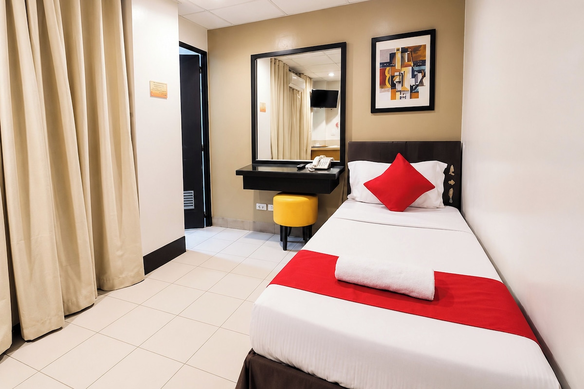 A hotel room with a bed, a bedside table, a mirror, a red pillow, and a yellow stool.