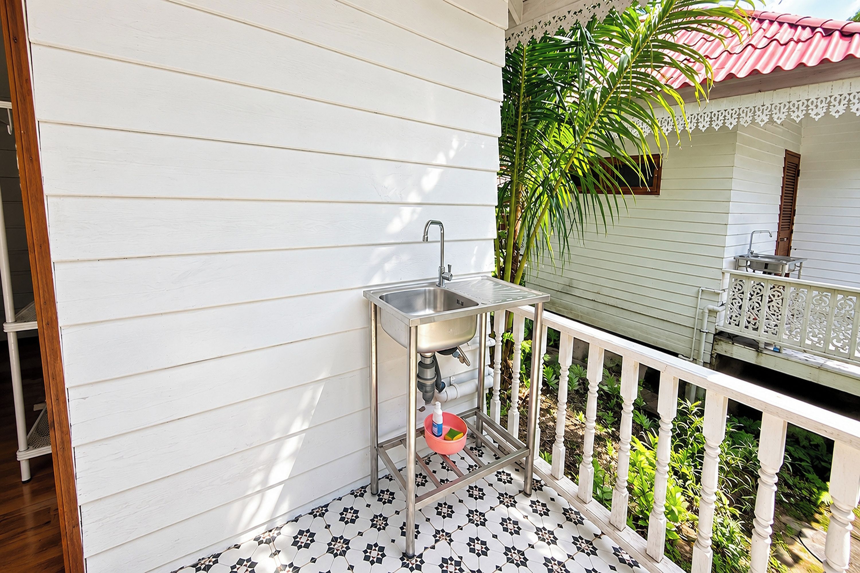 A small outdoor area with a metal sink and a white railing, surrounded by greenery and a tiled floor.
