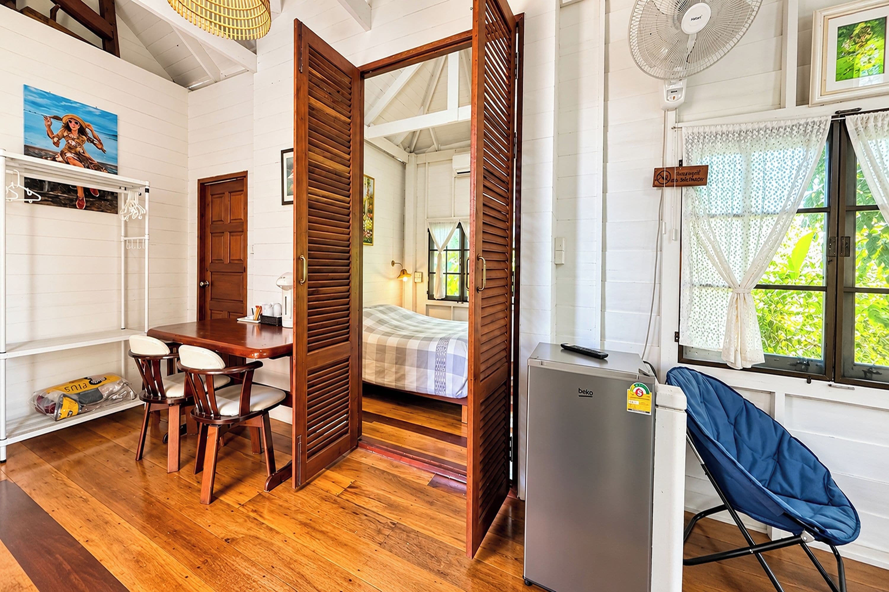 A room with a wooden floor, a dining table with chairs, a bed with a checkered bedspread, a refrigerator, and a window with a view of greenery.