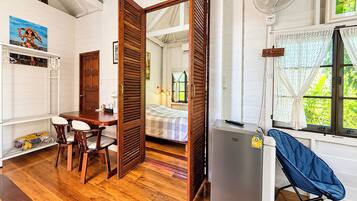 A room with a wooden floor, a dining table with chairs, a bed with a checkered bedspread, a refrigerator, and a window with a view of greenery.
