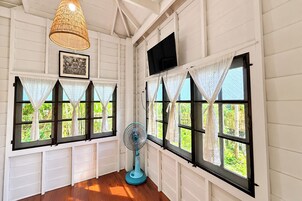 Una habitación con paredes de color blanco, paneles decorativos, ventilador de techo, televisor montado y ventanas con cortinas de encaje.