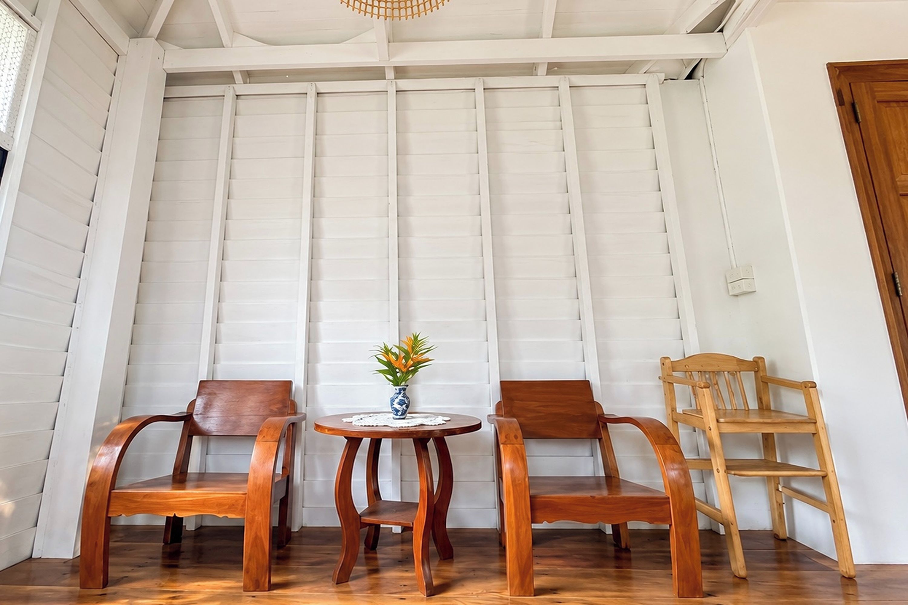 Una habitación con dos sillas de madera, una mesita con un florero y una silla de madera apoyada en una pared blanca con paneles.