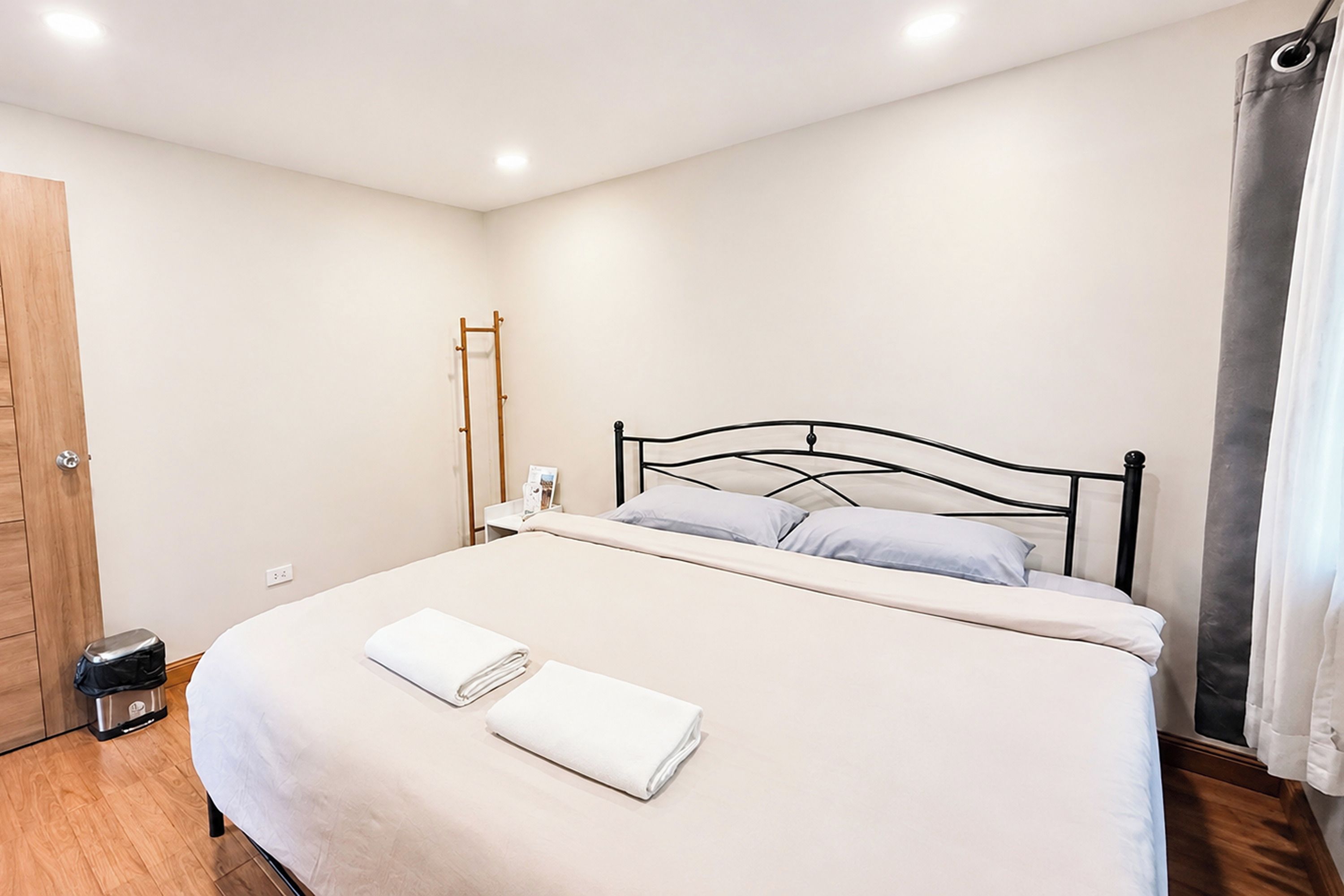Una cama bien hecha con sábanas blancas y dos almohadas, suelo de madera, una puerta y una lámpara fijada a la pared.