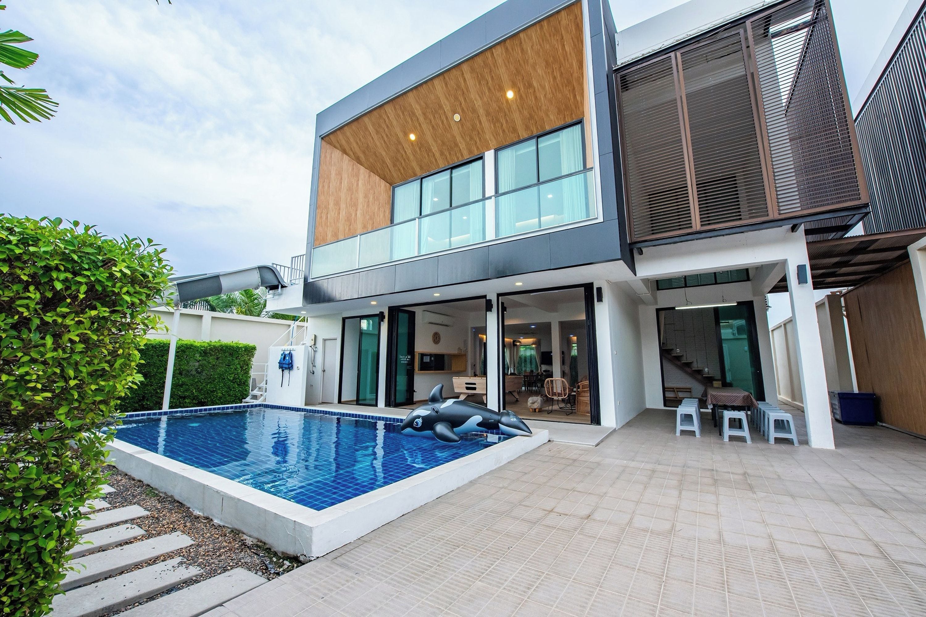 A modern two-story house with a swimming pool, outdoor seating, and a large window.