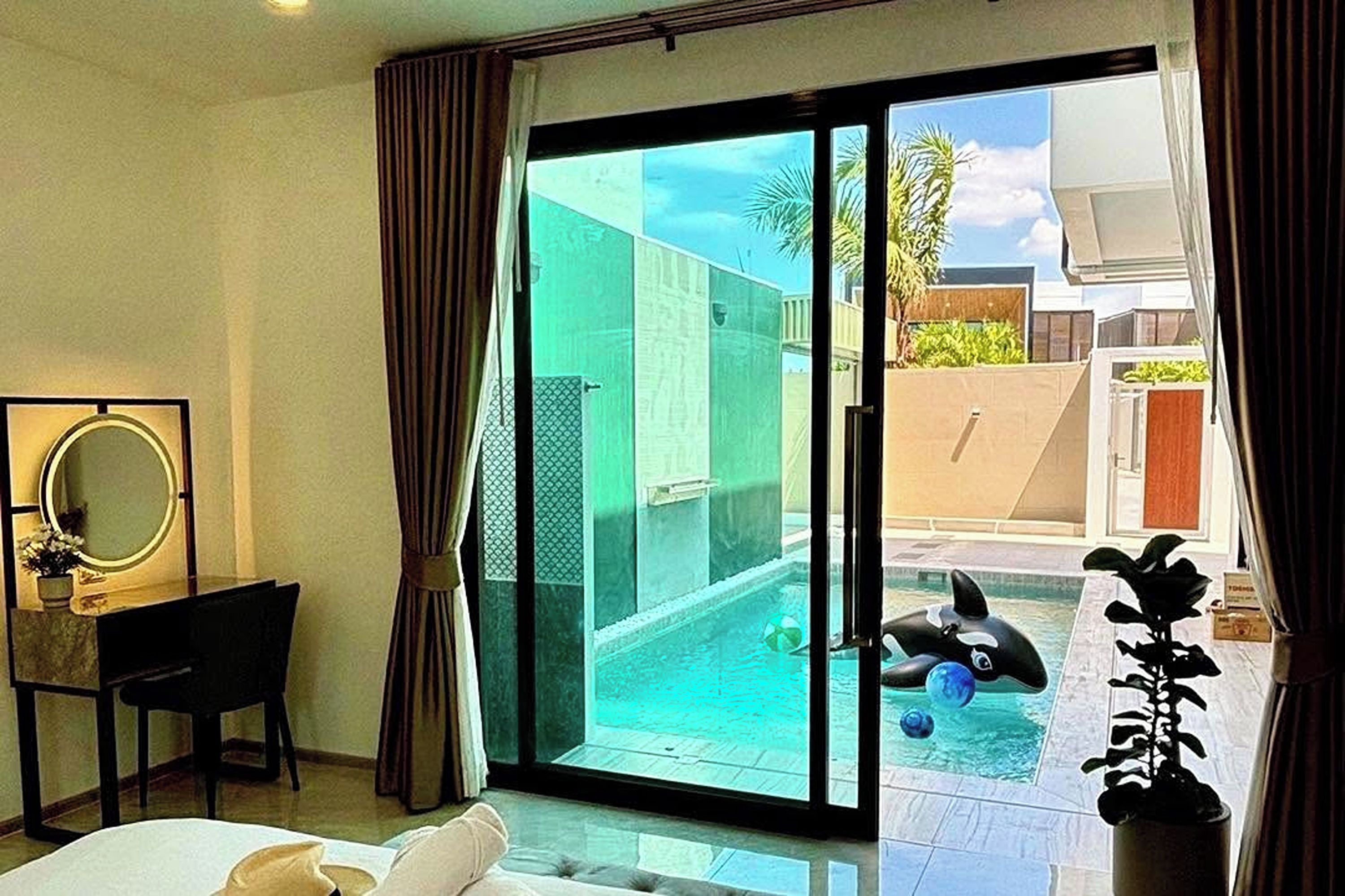 A hotel room with a bed, a desk, a chair, a large window with curtains, and a view of a swimming pool and outdoor area.