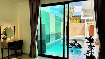 A hotel room with a bed, a desk, a chair, a large window with curtains, and a view of a swimming pool and outdoor area.