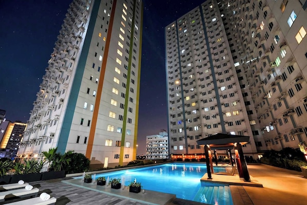 Vista nocturna de un edificio residencial de gran altura con piscina y zona de descanso.
