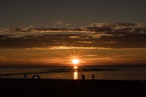 Ein Sonnenuntergang über einem Strand, im Vordergrund sind Silhouetten von Menschen und eine Struktur zu sehen.