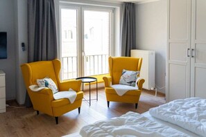A room with two yellow armchairs, a small round table, a bed with white bedding, and a view through a window with grey curtains. - House Panorama WHG 33 (Cuxhaven)