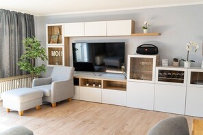 A modern living room with a flat-screen TV, a grey sofa, a small ottoman, and built-in shelving with decorative items and a radiator. - House Panorama WHG 33 (Cuxhaven)
