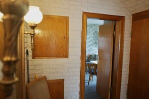 Un couloir avec une porte en bois, une lampe fixée au mur, et un aperçu d’un coin repas avec des chaises et des tables.