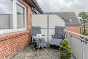 Ein Balkon mit Tisch und zwei Stühlen, einem kleinen Topfpflanzen und Blick auf ein Backsteinhaus.