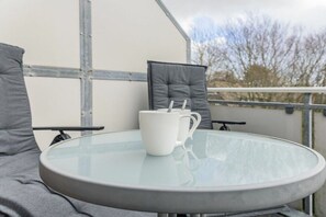 Une table ronde en verre, sur laquelle sont posés un mug blanc et une cuillère, le tout placé devant un fauteuil gris rembourré et un mur gris.