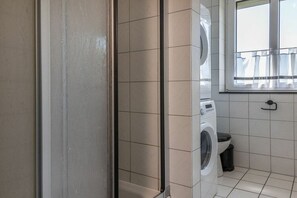 Een praktische wasruimte met een wasmachine, een klein afvalbakje en een raam met gordijnen.