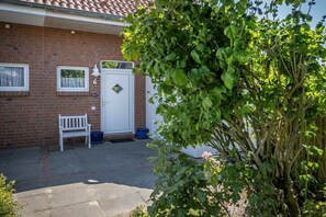 Een bakstenen huis met een witte deur, een bankje en een boom op de voorgrond.