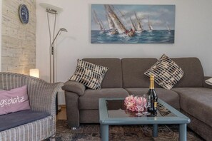 A living room with a sofa, a coffee table with a bottle and glasses, a painting of sailboats, and a patterned pillow. - 50006 House Deichkieker (Harlesiel)