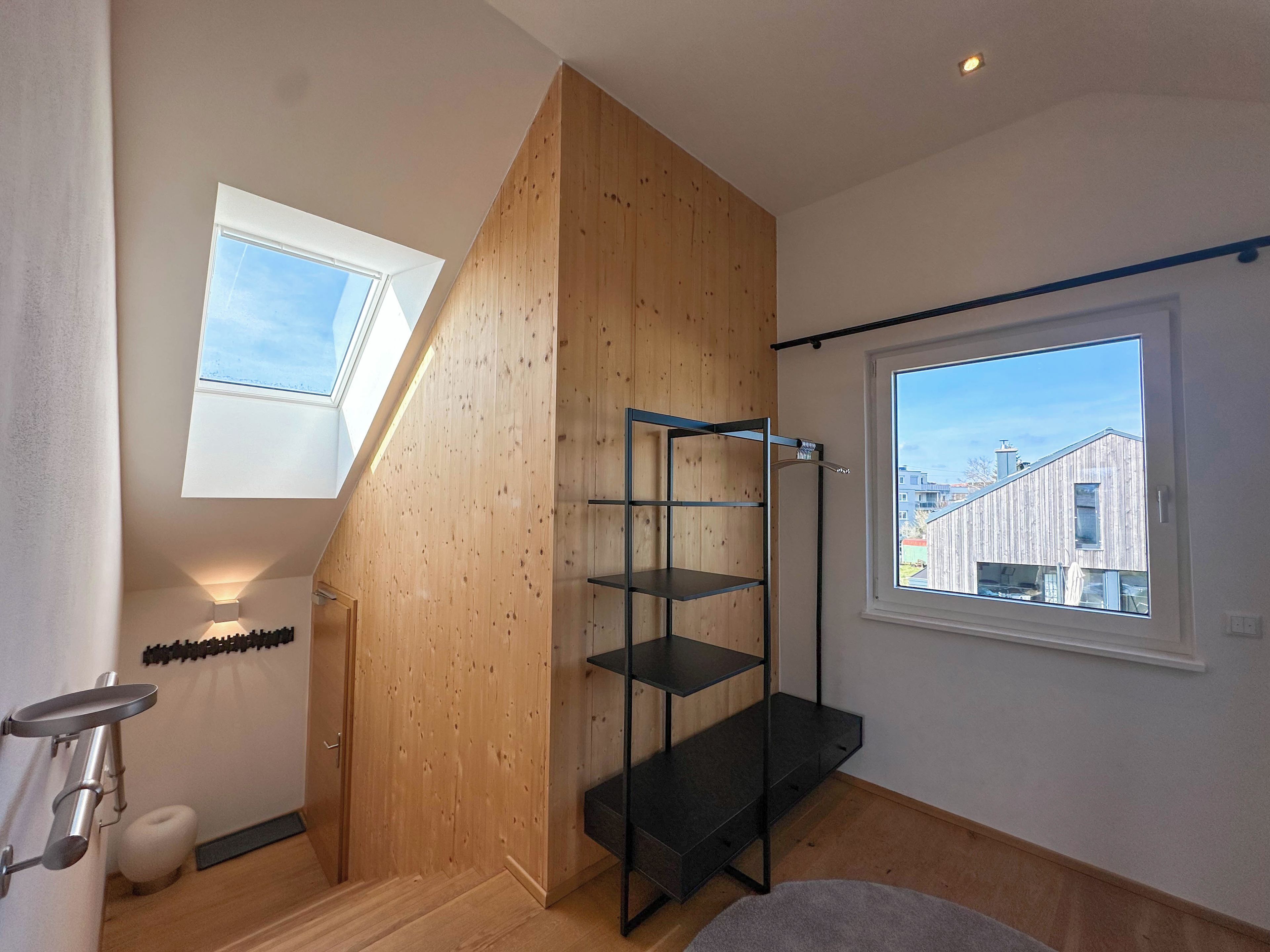 A room with a wooden sloped ceiling, a black metal shelf unit, a window with a view of buildings, and a small round stool.