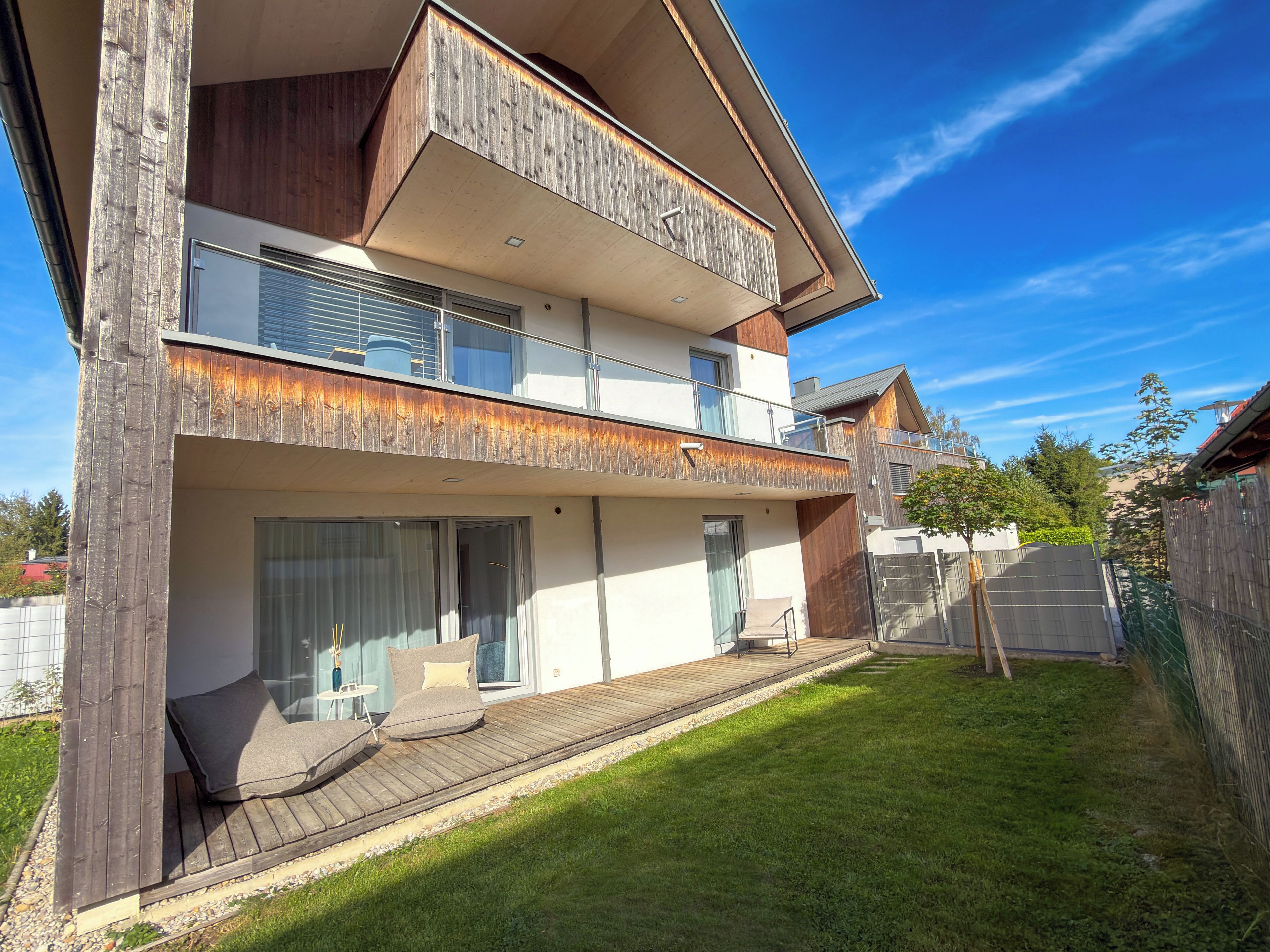 Ein modernes Wohnhaus mit zwei Etagen, einem Balkon, einer Holzterrasse und einem gepflegten Rasen.
